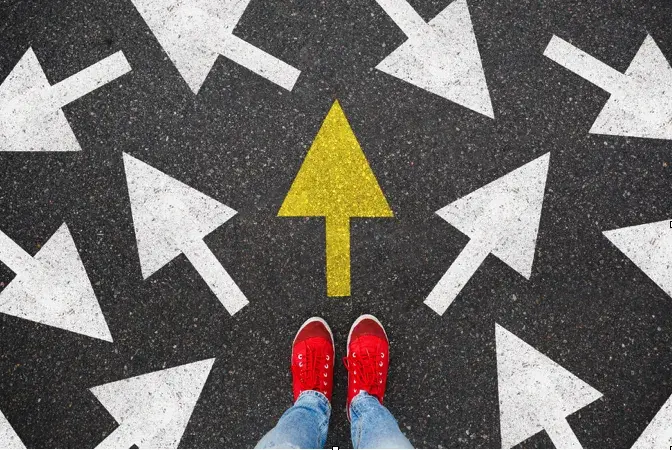 A view of jeans and red sneakers standing in front of several white arrows on asphalt pointing in different directions with a yellow arrow point forward. 